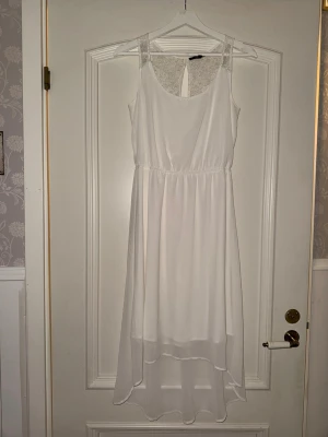 Vit långklänning med spetsdetaljer - Elegant vit långklänning med ärmlös design och vackra spetsdetaljer upptill. Klänningen har rund halsringning, lätt markerad midja och lätt material. Perfekt för sommaren, fest och student.