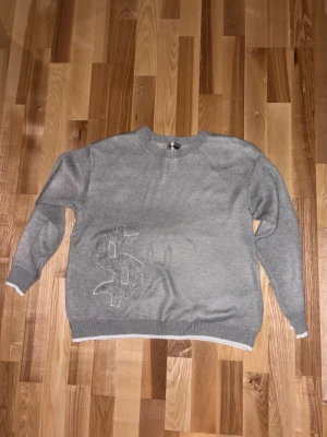 Dollar tröja  - Grå stickad tröja med vit kant vid ärmslut och nederkant. På framsidan finns ett stort, diskret dollartecken och på ryggen står det 'buy happiness' i vit text. Tröjan har rund hals och lång ärm, perfekt för en avslappnad streetwear-look. 