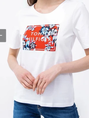 Vit T-shirt Tommy Hilfiger blommig - Vit T-shirt från Tommy Hilfiger med korta ärmar och rund hals. På bröstet finns en stor röd patch med blommigt mönster i blått, vitt och svart samt varumärkets logga. Tillverkad i mjuk bomull, perfekt för en avslappnad och trendig look.