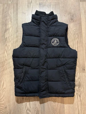 Abercrombie & Fitch - Snygg svart dunväst från Abercrombie & Fitch med hög krage och två fickor framtill. Västen har ett broderat logomärke på bröstet och quiltad design. Perfekt för lager-på-lager och streetstyle.
