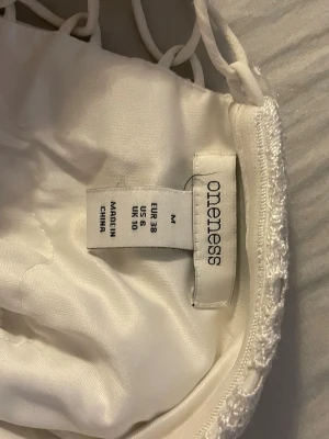 Vit spetsklänning från Oneness - Vit klänning från Oneness med fina spetsdetaljer och snörning i midjan. Klänningen har en elegant look med dekorativa spetsmönster längs sidorna och är tillverkad i ett mjukt tyg som känns lyxigt mot huden.