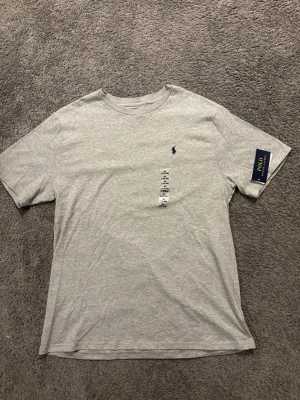Grå t-shirt från Polo Ralph Lauren - Snygg grå t-shirt från Polo Ralph Lauren med klassisk passform och rund hals i storlek XL. Tshirten är aldrig använd och har kvar prislapp från köpet och är därav i nyskick. Skriv gärna vid frågor och funderingar!😃✅