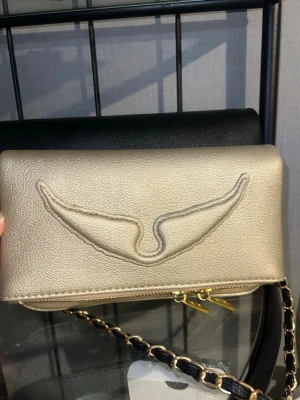 Guldig plånbok med kedja - Snygg guldig plånbok med kedjedetalj och dragkedja i guld. Plånboken har ett broderat vingliknande mönster på framsidan och är tillverkad i ett glansigt konstlädermaterial. Perfekt för dig som vill ha en trendig accessoar med plats för det viktigaste. Ny oanvänd fick i student present, nypris 3490