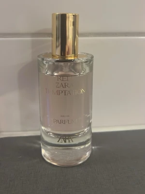 Zara Red Temptation Eau de Parfum - Zara Red Temptation Eau de Parfum i en elegant, rund glasflaska med guldfärgat lock. Flaskan har en minimalistisk design med guldtext och transparent glas som visar den ljusrosa vätskan inuti. Perfekt för dig som gillar lyxiga och moderna dofter.