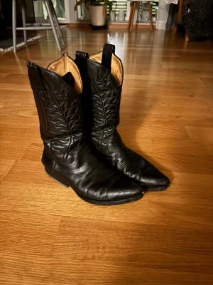 Tony mora vintage boots  - Säljer ett par svarta cowboy boots i läder med spetsig tå och dekorativa sömmar längs skaftet. Insidan är ljust beige och bootsen har dragflikar upptill för enkel påtagning. Perfekta för dig som vill ha en tidlös westernstil.