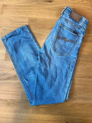 Blå jeans från Nudie - Säljer ett par klassiska blå jeans från Nudie Jeans med raka ben och snygga kontrastsömmar. Jeansen har den ikoniska broderade vågen på bakfickorna och är gjorda i mjuk denim. Perfekta för en avslappnad och trendig look.