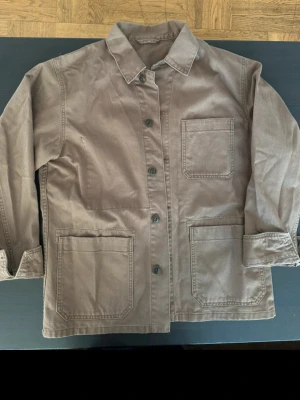 Brun overshirt med stora fickor - Säljer en stilren brun overshirt i kraftigt bomullstyg. Jackan har tre stora utanpåliggande fickor framtill, knäppning med knappar och klassisk krage. Perfekt för lager på lager och enkel att matcha med olika outfits.