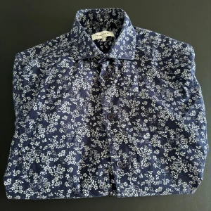 Blommig blå skjorta från Studio Total - Snygg blå skjorta från Studio Total med vitt blommigt mönster. Skjortan har klassisk krage.