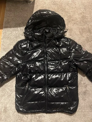 Svart glansig dunjacka från moncler - Snygg svart dunjacka från Moncler med glansigt yttermaterial och vadderad design. Jackan har huva, två dragkedjefickor framtill och Moncler-logga på ärmen. Perfekt för kalla vinterdagar och ger en trendig look. Rep!
