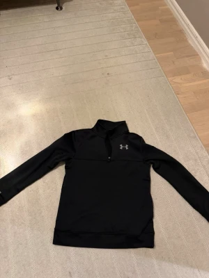 Svart träningströja från Under Armour - Svart långärmad träningströja från Under Armour med halv dragkedja och hög krage. Tröjan har en diskret logga på bröstet och är tillverkad i ett stretchigt funktionsmaterial som passar perfekt för träning och sport.