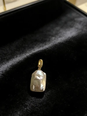 Halsbandshänge med barockpärla och guld - Unikt halsbandshänge med en stor, oregelbunden barockpärla i vit skimrande nyans. Hänget har ett guldfärgat fäste med diskret mönster, vilket ger en cool och trendig look. Perfekt för dig som gillar statement-smycken och vill sticka ut.