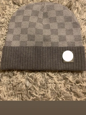 Grå rutmönstrad mössa med patch - Säljer en stilren grå mössa med rutmönster och vikt kant. Mössan har en ribbstickad kant och en fyrkantig patch framtill. Perfekt accessoar för kalla dagar och passar till de flesta outfits.