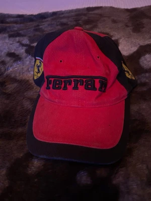 Röd och svart Ferrari keps - Snygg röd och svart keps från Ferrari med broderad logga på sidan och texten 'Ferrari' framtill. Klassisk böjd skärm och justerbar passform. Perfekt för dig som gillar motorsport och vill ha en ikonisk accessoar.