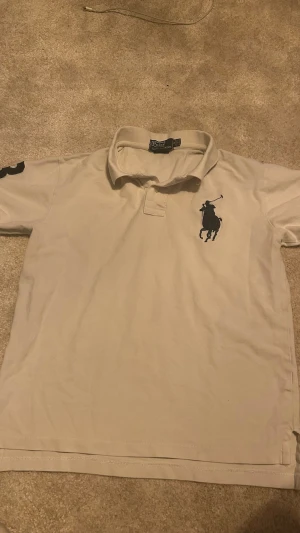 Vit pikétröja från Polo Ralph Lauren - Klassisk vit pikétröja från Polo Ralph Lauren med stor svart logga på bröstet och krage. Tröjan har korta ärmar och knappar vid halsen. Perfekt för en clean och stilren look.