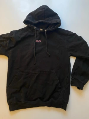 Svart hoodie från SNUSK med tryck - Svart hoodie från SNUSK med huva och dragsnören. Liten SNUSK-logga i rosa på bröstet och stort tryck av en ryttare på häst i rosa på ryggen. Klassisk känguruficka framtill. Perfekt för dig som gillar streetwear och unika prints.