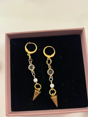 Guldiga örhängen med pärlor och pikar - Säljer ett par unika guldiga örhängen med små vita pärlor, transparenta stenar och coola konformade hängen. Örhängena har en modern design med ringar och detaljer i metall. Perfekta för dig som vill sticka ut lite extra.