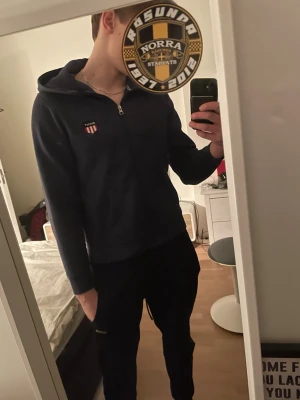 Mörkblå hoodie från GANT med dragkedja - Säljer en mörkblå hoodie från GANT med dragkedja och huva. Hoodien har ett broderat GANT-märke på bröstet med röd och gul sköld. Insidan är mjuk och fleecefodrad för extra komfort. Perfekt för dig som gillar stilrena och klassiska plagg. Ganska andvänd. Inga frågår, bara prisförslag. Snabb köp!