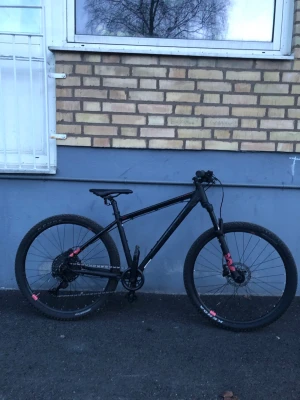 Svart mountainbike med rosa detaljer - Säljer en svart mountainbike med mattsvart ram och rosa detaljer på hjulen och framgaffeln. Cykeln har grova däck, skivbromsar och sportig design. Perfekt för dig som gillar att cykla i skogen eller på grusvägar. Cykeln är god skick (har ej pumpa hemma så jag kan inte pumpa cykeln)