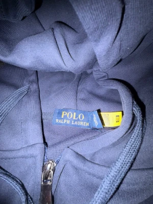Marinblå hoodie från Polo Ralph Lauren - Snygg marinblå hoodie från Polo Ralph Lauren med dragkedja framtill och klassisk huva. Hoodien har fickor på magen och den ikoniska broderade loggan på bröstet. Perfekt för en chill och stilren look. Materialet känns mjukt och bekvämt.
