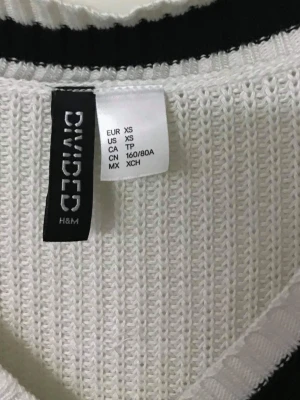 Vit stickad tröja med svarta ränder - Snygg vit stickad tröja från H&M Divided med svarta ränder på ärmarna, vid halsringningen och nederkanten. Tröjan har V-ringning och lång ärm, perfekt för en avslappnad och trendig look.