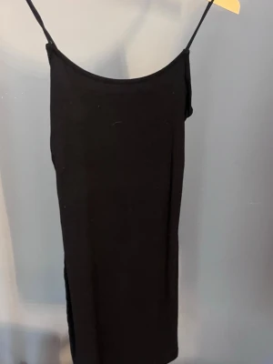 Svart långklänning med smala band - En enkel och stilren svart långklänning med smala axelband och rak passform. Klänningen har en slits på sidan och är ärmlös, vilket ger en clean och modern look. Perfekt att styla med sneakers eller klackar.