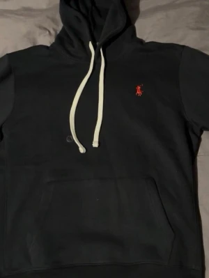 Svart hoodie från Ralph Lauren - Snygg svart hoodie från Ralph Lauren med vit dragsko och klassisk röd logga på bröstet. Hoodien har en stor känguruficka framtill och är tillverkad i mjukt material som passar perfekt för en chill och stilren look.
