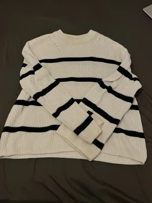Vit stickad tröja med svarta ränder - Säljer en vit stickad tröja med svarta horisontella ränder. Tröjan har rund halsringning, långa ärmar och är ribbstickad för en skön och avslappnad look. Perfekt för dig som gillar klassiska och stilrena plagg.