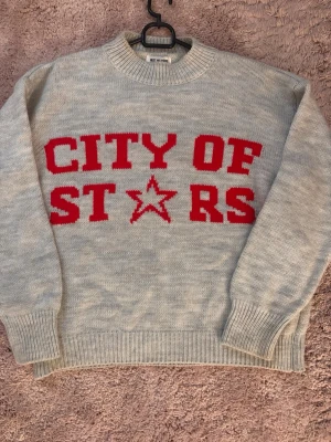 Grå stickad tröja med röd text - Stickad tröja i ljusgrått med stor röd text 'CITY OF STARS' och en stjärna framtill. Tröjan har rund halsringning, ribbade muddar och avslut. Perfekt för dig som gillar statementplagg och vill ha en mysig, trendig look.