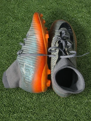 Nike CR7 grå-orange fotbollsskor - Snygga fotbollsskor från Nike CR7-kollektionen i grått med orange sula och svarta detaljer. Skorna har strumpliknande skaft för extra stöd och snörning framtill. Perfekta för dig som vill sticka ut på planen med både stil och funktion.