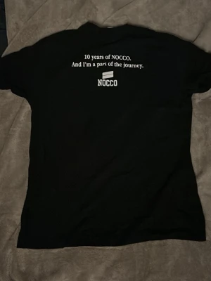 Svart NOCCO t-shirt med tryck - Säljer en svart t-shirt från NOCCO med tryck både fram och bak. På bröstet står NOCCO-loggan och på ryggen står texten '10 years of NOCCO. And I'm a part of the journey.' Klassisk passform och rund hals.