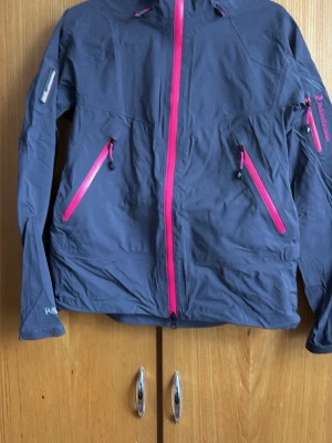 Peak Performance vindjacka mörkgrå/rosa - Snygg mörkgrå vindjacka från Peak Performance med rosa dragkedjor och detaljer. Jackan har huva, flera fickor med dragkedja och logga på ärmen. Perfekt för blåsiga dagar och outdoor-aktiviteter.