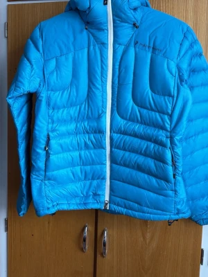 Blå dunjacka från Peak Performance - Snygg blå dunjacka från Peak Performance med huva och vit dragkedja. Jackan har två sidofickor med dragkedja och diskret logga på bröstet. Perfekt för kalla vinterdagar tack vare det värmande dunmaterialet och den sportiga designen.