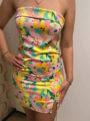 Blommig axelbandslös kortklänning - Färgglad axelbandslös kortklänning med blommigt mönster i gult, rosa, grönt och blått. Klänningen har en figurnära passform, liten slits framtill och är tillverkad i ett glansigt syntetmaterial. Perfekt för dig som vill sticka ut med en somrig och lekfull look.