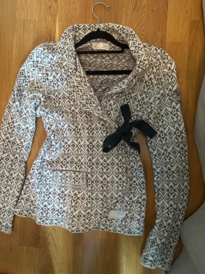 Mönstrad kofta med rosett Odd Molly - Säljer en snygg kofta från Odd Molly med gråvitt mönster, lång ärm och dekorativ rosett framtill. Koftan har broderad detalj på ärmen och en skön, stickad känsla. Perfekt för dig som gillar unika detaljer och mysiga plagg.