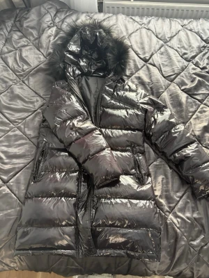 Glansig svart dunjacka med pälshuva - Säljer en lång, glansig svart dunjacka med äkta päls. Jackan har två dragkedjeförsedda fickor och quiltad design som ger en puffig look. Perfekt för kalla vinterdagar. Trasig i ena fickan men går att sy.