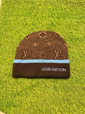 Brun Louis Vuitton mössa med logga - Säljer en brun mössa från Louis Vuitton med klassiskt monogrammönster och ljusblå kant. Mössan har broderad Louis Vuitton-text framtill och är tillverkad i mjukt stickat material. Perfekt accessoar för dig som vill ha en lyxig och trendig look.
