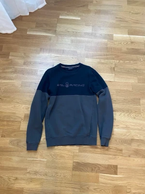 Sail Racing tröja - Snygg sweatshirt från Sail Racing i grått och marinblått med logga broderad på bröstet. Tröjan har rund halsringning, långa ärmar och ribbade muddar vid ärmslut och nederkant. Perfekt för en avslappnad och sportig stil.