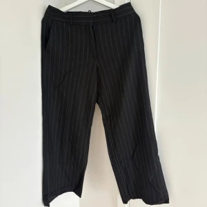 Svarta kostymbyxor med kritstreck STOCKH LM - Snygga svarta kostymbyxor från STOCKH LM med vita kritstrecksränder. Byxorna har raka ben, bälteshällor och sidofickor. Perfekta för en stilren och trendig look. Materialet känns mjukt och har en klassisk kostymkänsla.