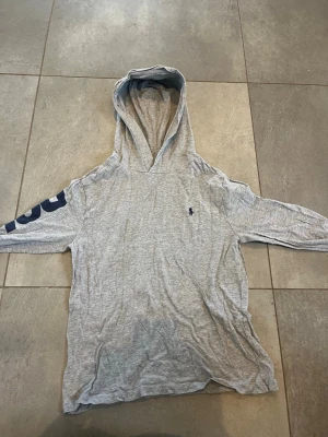 Grå hoodie från Polo Ralph Lauren - Säljer en grå hoodie från Polo Ralph Lauren med blå logga broderad på bröstet och tryck på ena ärmen. Hoodien har en avslappnad passform och är tillverkad i mjukt bomullsmaterial. Perfekt för en chill och sportig look.
