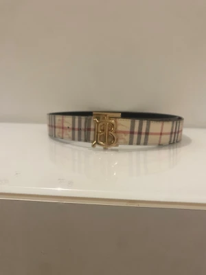 Burberry bälte med guldspänne - Snyggt Burberry bälte med klassiskt rutmönster i beige, svart och rött. Bältet har ett stilrent guldigt 'TB'-spänne framtill och insidan är svart. Perfekt accessoar för att lyfta din outfit.