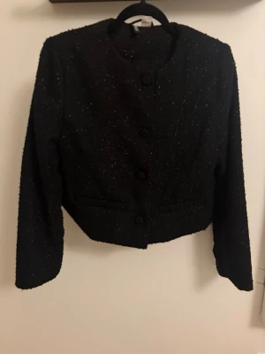 Svart kavaj från H&M med glitter - Stilren svart kavaj från H&M med diskret glittereffekt i tyget. Rak, kortare modell med rund halsringning och stora klädda knappar framtill. Två dekorativa fickor och lång ärm. Perfekt för dig som vill ha en chic och trendig look.