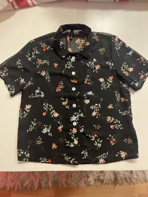 Svart blommig skjorta med knappar - Kortärmad svart skjorta med blommigt mönster. Skjortan har vita knappar framtill. Materialet känns lätt och luftigt.