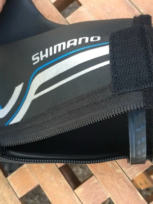 Shimano svarta skoöverdrag S2000D - Säljer ett par svarta skoöverdrag från Shimano, modell S2000D. De är tillverkade i neopren med detaljer i blått, vitt och silver samt har kardborreknäppning. Perfekta för att skydda skorna mot regn och vind under cykling. Strl 43