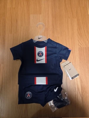 Nike PSG Barn Set 3-6 månader, 65-70 cm - Ett sött matchande set för barn från säsong 22/23 , bestående av en mörkblå tröja med ett vertikalt band i vitt och rött på framsidan, prydd med Paris Saint-Germains emblem och Nike-loggan. Setet inkluderar även ett par matchande mörkblå shorts med Nike-loggan samt strumpor.  65-70 cm