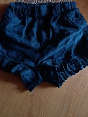 Mörkblå volangshorts med resår - Supersöta mörkblå shorts med volangdetaljer längs bensluten och bekväm resår i midjan. Shortsen har fickor både fram och bak och är perfekta för varma dagar. De är tillverkade i ett mjukt och lätt material som känns skönt mot huden.