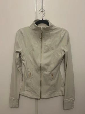 Lululemon define jacket - Snygg vit träningsjacka med figurnära passform. Jackan har guldiga dragkedjor framtill och på fickorna. Perfekt för när man vill se sportig ut eller vardaglig. Färgen är krämvit. Storlek 4 motsvarar XS / S 