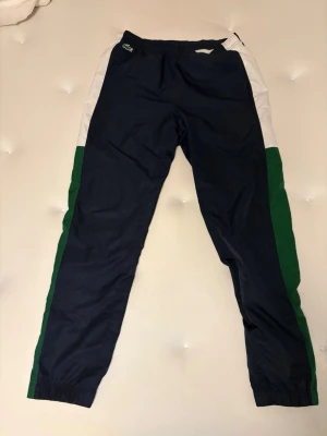 Lacoste trackpants  - Säljer ett par Lacoste-byxor i väldigt fint skick och sparsamt använda. Färg: blå, grön och vit Storlek: FR 3 (motsvarar S, men passar även M beroende på fit) Skick: 9/10 – inga skador eller slitningar Byxorna har dragkedjor längst ner vid benen så du kan justera passformen och välja hur vida du vill ha dem. Stilrena och bekväma med riktigt snygg detaljering.