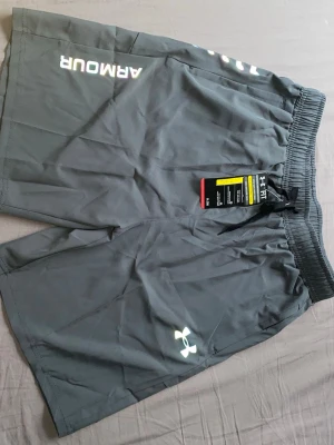 Grå träningsshorts från Under Armour - Snygga grå träningsshorts från Under Armour med elastisk midja och logga på benet. Lätta och bekväma shorts perfekta för gymmet eller löprundan. Klassisk design med tryckt text och logotyp i vitt.
