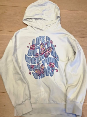 Vit hoodie med fjärilar och text - Vit hoodie med tryck i pastellfärger framtill. Motivet består av lila texten 'Life is better when you wear your wings' samt rosa och gula fjärilar och blommor. Hoodien har huva och ribbade muddar vid ärmslut och nederkant. Perfekt för en färgglad och lekfull stil.