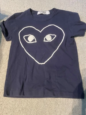 Mörkblå Comme des Garçons Play t-shirt - Mörkblå t-shirt från Comme des Garçons Play med det ikoniska hjärtmotivet med ögon i vitt på bröstet. Klassisk rund hals och korta ärmar. Tillverkad i mjuk bomull som är skön att bära. Storlek S men skulle säga att den passar mer som en XS.
